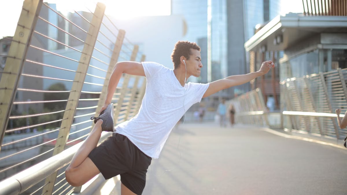 Determined ethnic sportsman in earphones sport clothes stretching legs while listening to music and training alone on street against blurred urban environment